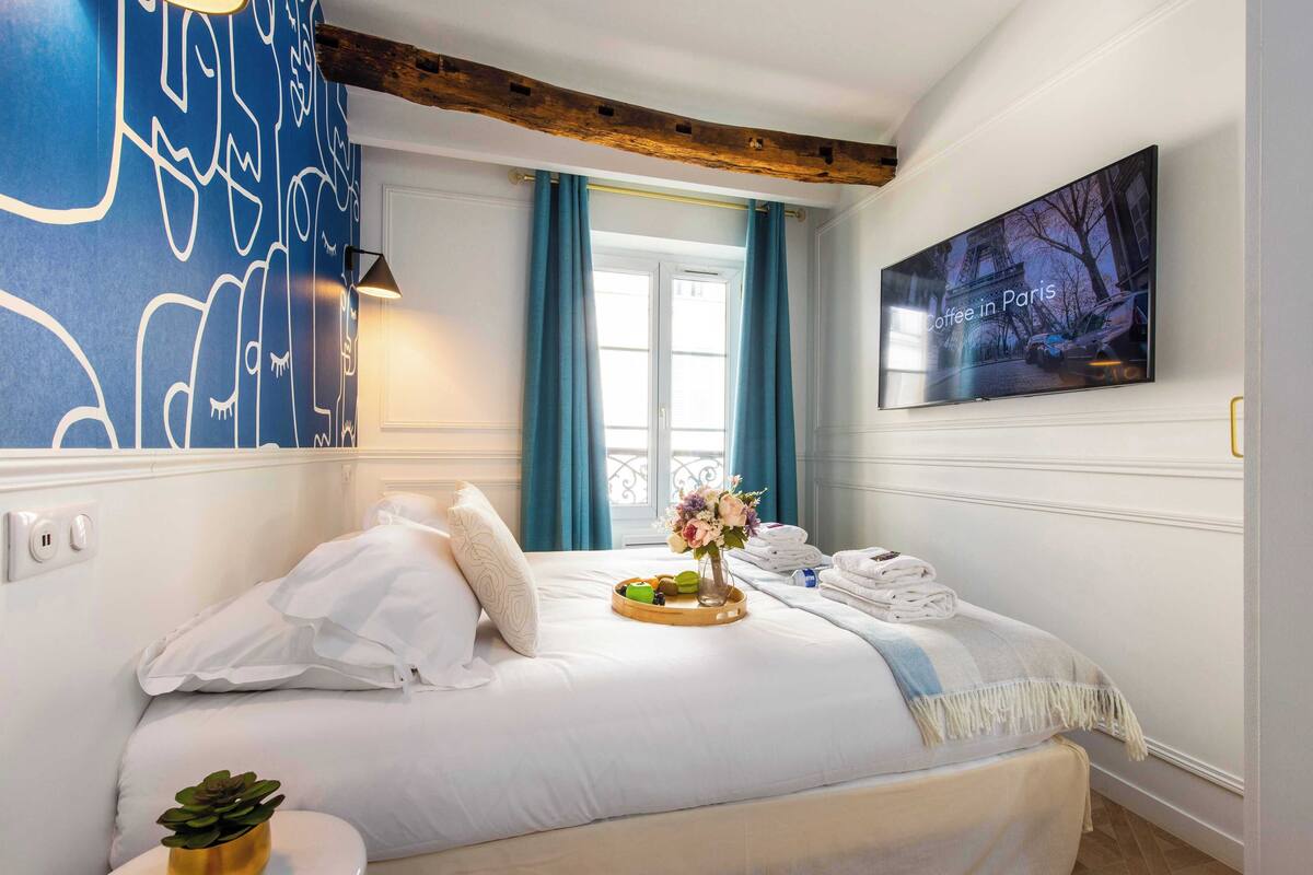 Quarto de hotel com cama, televisão na parede, janela com cortinas e mesa de cabeceira com uma planta.