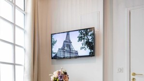 Habitación de esquina con una mesa redonda, dos otomanas, un televisor en la pared y una ventana con cortinas.
