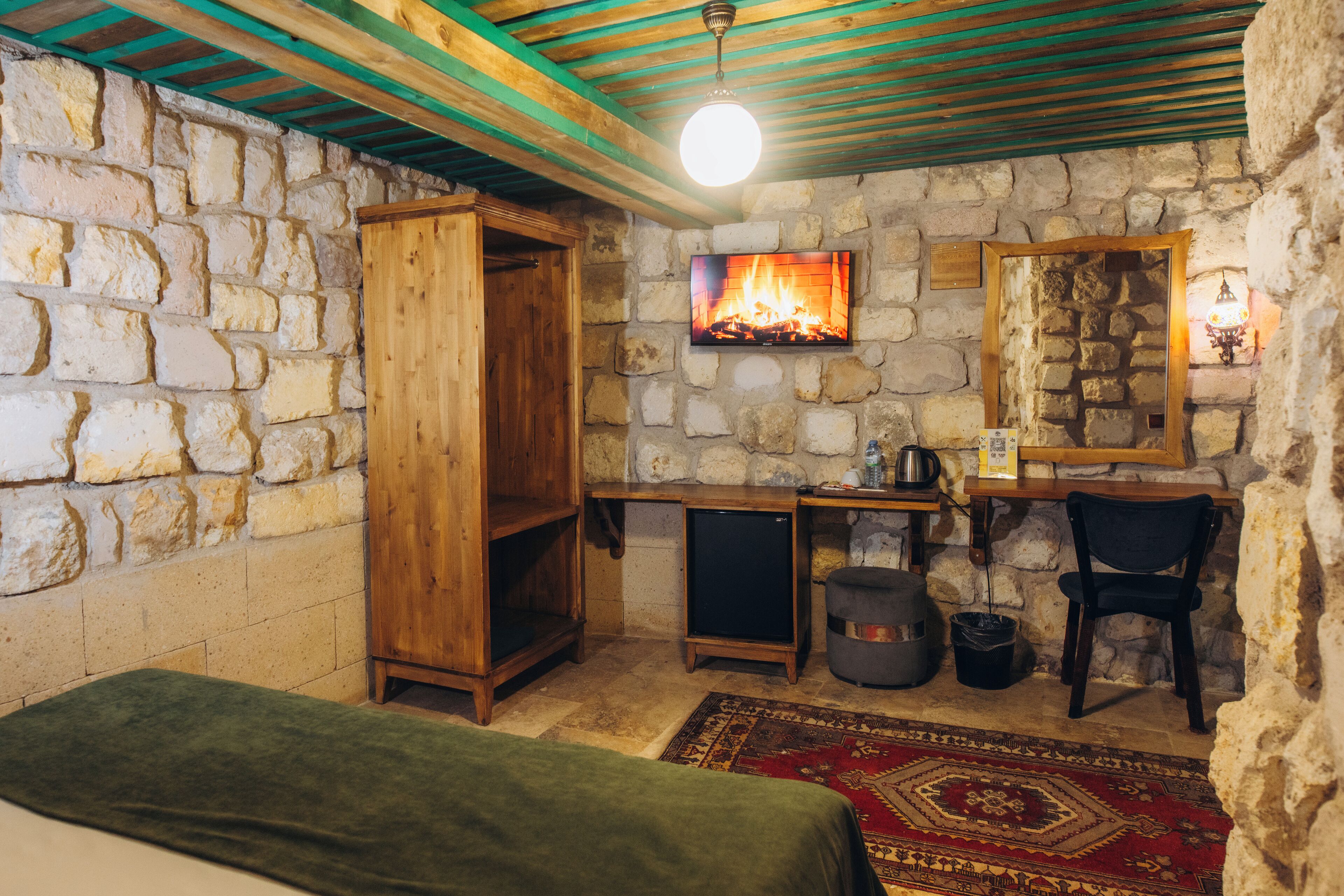 A rustic room with stone walls, a wooden wardrobe, a television mounted on the wall, a chair, a small table with a kettle, and a red patterned rug.