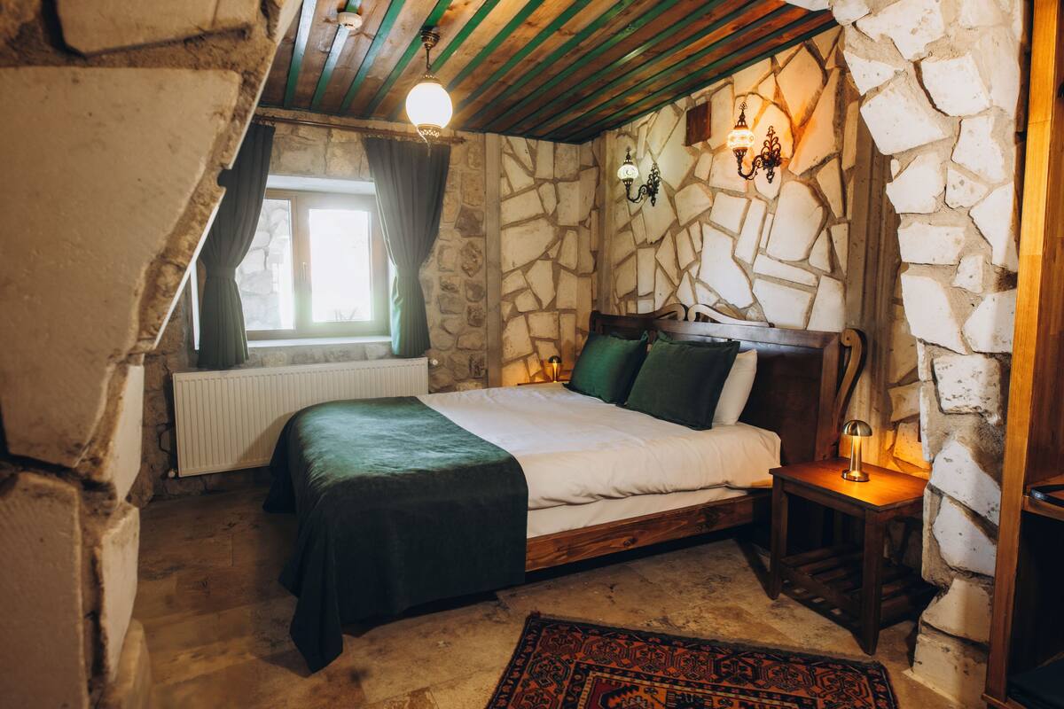 Une chambre à coucher rustique avec un mur en pierre, un cadre de lit en bois, une table de chevet et une fenêtre avec des rideaux.