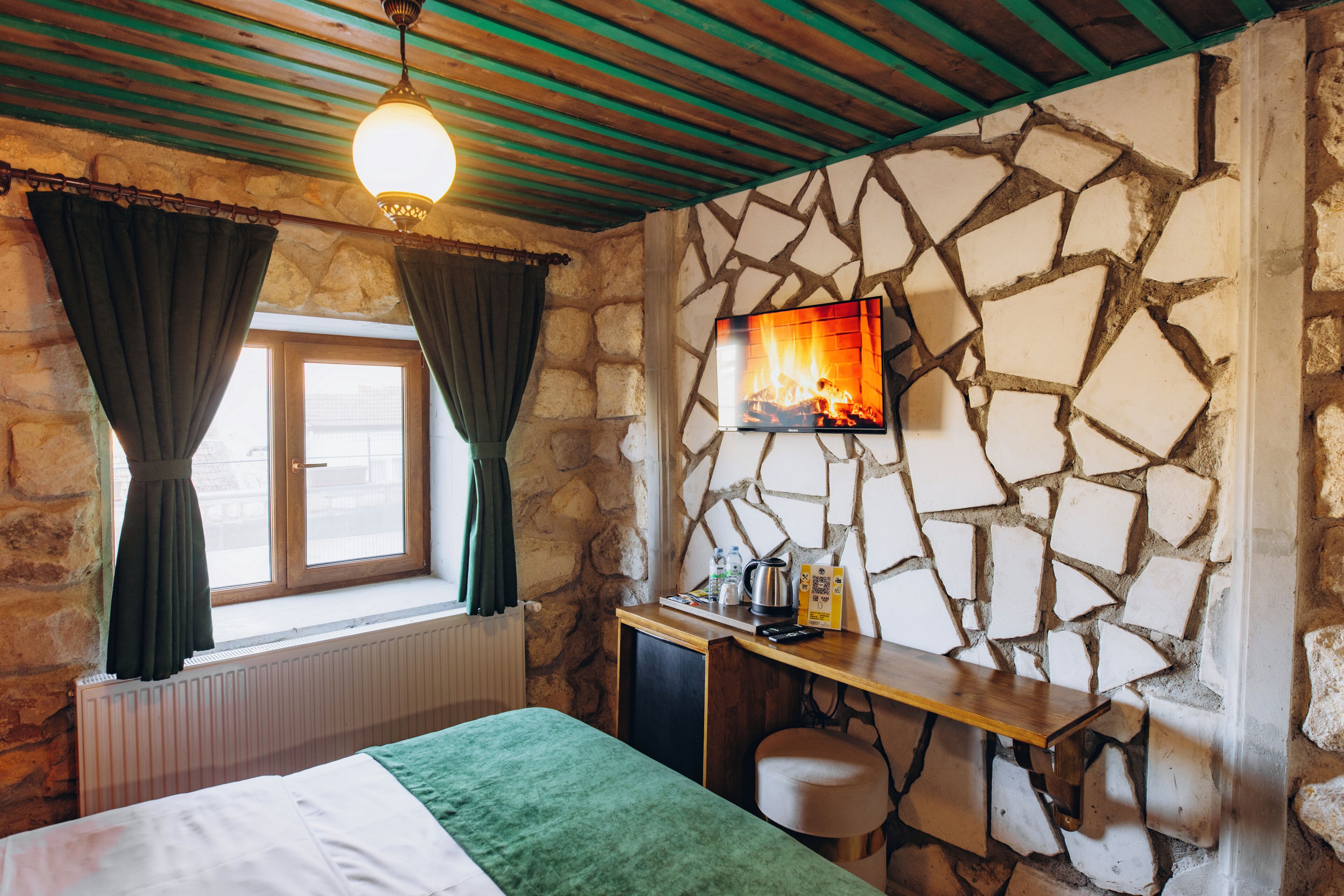 A cozy room with a stone wall, a bed, a desk, and a TV displaying a fireplace.