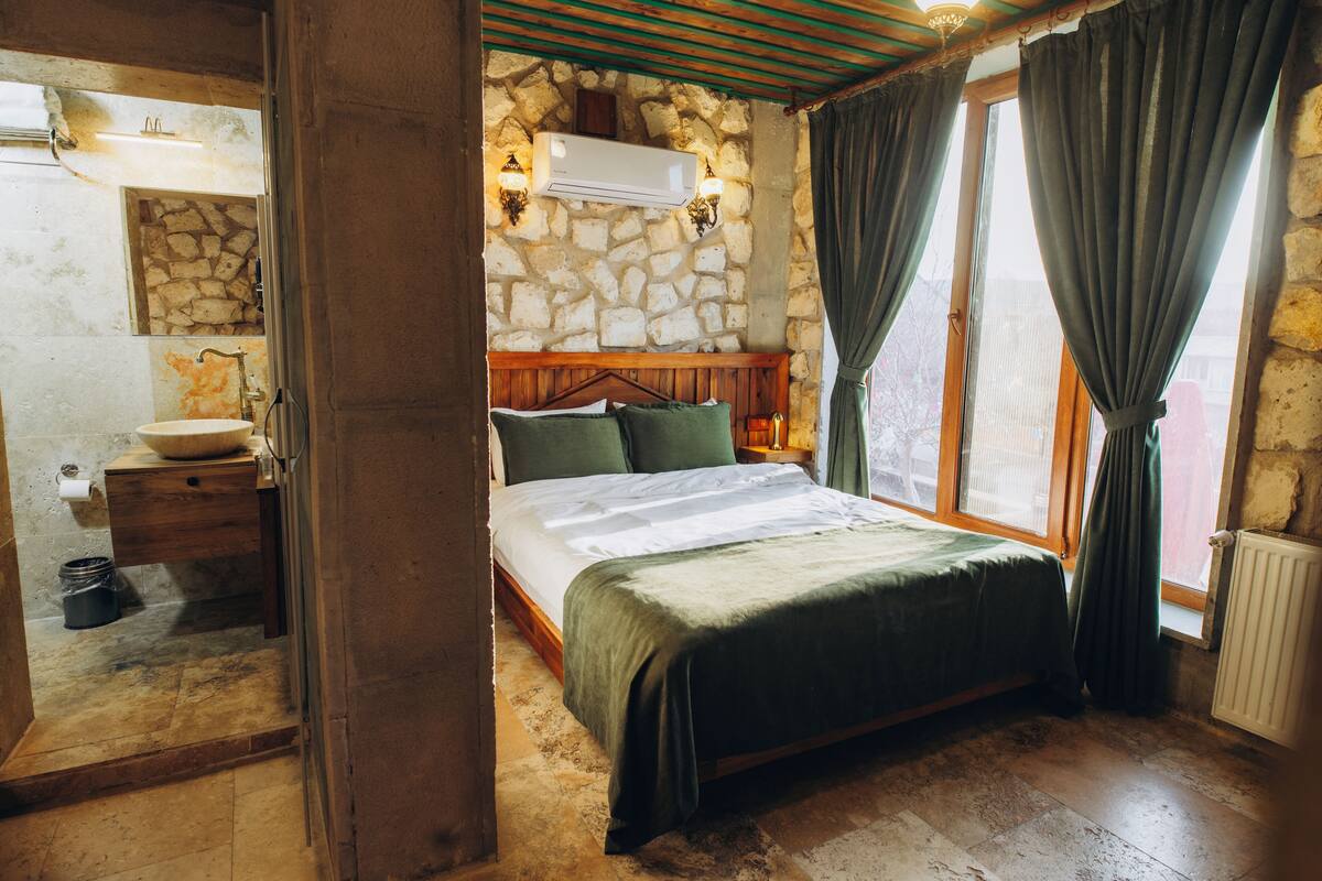 Une chambre avec un mur en pierre, un lit en bois, une table de chevet, un lavabo et une fenêtre avec des rideaux.