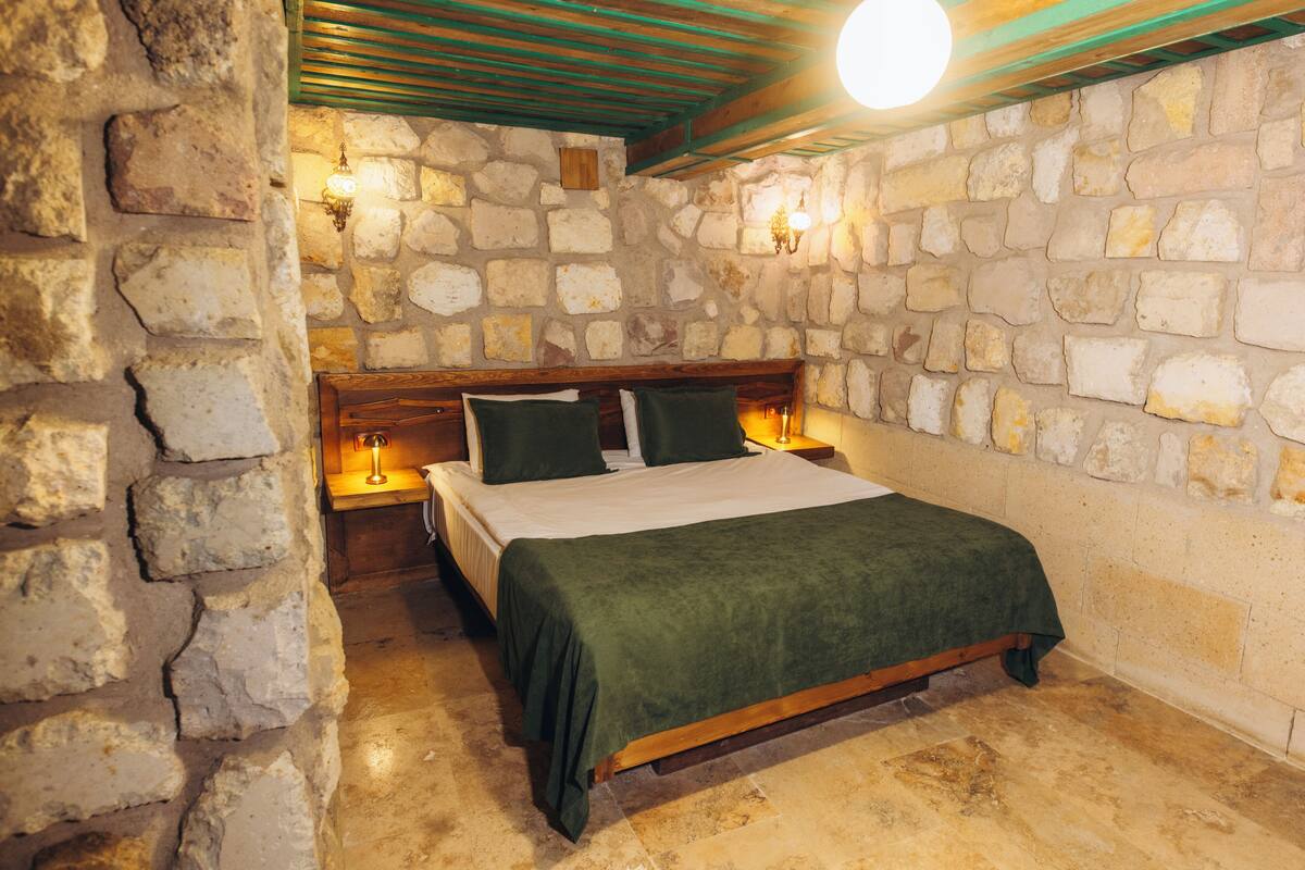 Une chambre aux murs de pierre, avec un lit en bois, une literie verte et deux lampes fixées au mur.