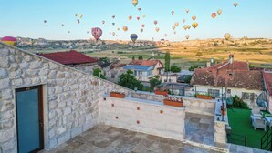 Una terraza en la azotea con muros de piedra, techo rojo y vistas a un pueblo con globos aerostáticos a lo lejos.