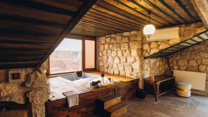 A rustic stone-walled bathroom with a wooden bathtub, a wooden bench, and a stone sink.
