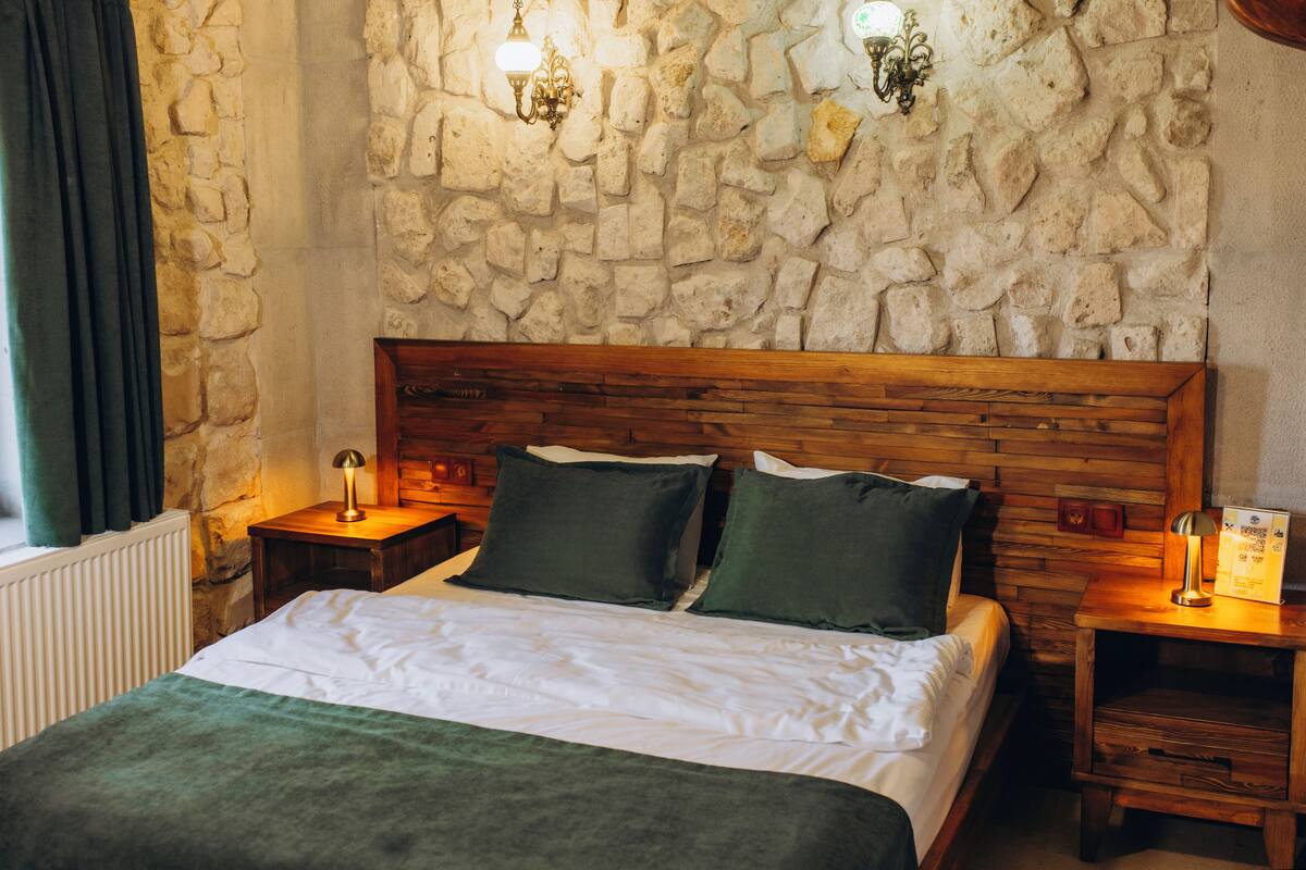 Une chambre avec un mur en pierre, une tête de lit en bois, un lit avec une literie blanche, deux coussins verts et deux tables de chevet avec des lampes.