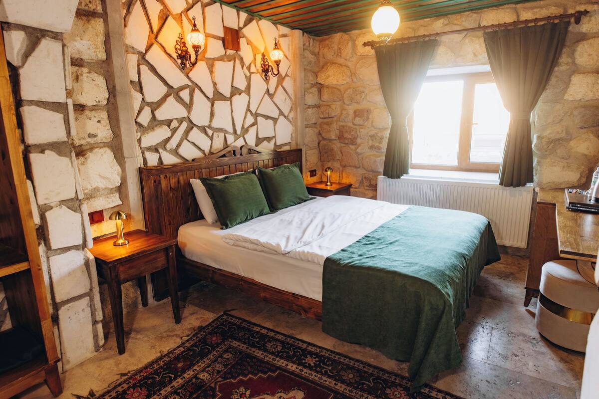 Une pièce avec un mur en pierre, un lit avec une literie verte, une table de chevet avec une lampe et une fenêtre avec des rideaux.