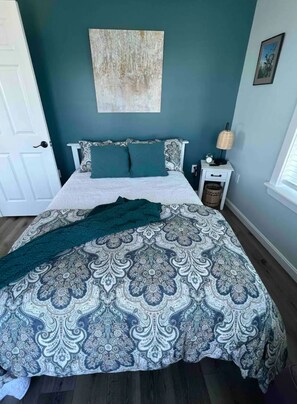 Un dormitorio con cama, mesita de noche, lámpara y una obra de arte en la pared.