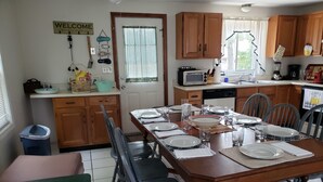 Comedor con una mesa de madera dispuesta para cuatro personas, rodeada de sillas. Se observa una zona de cocina con armarios de madera y un microondas.