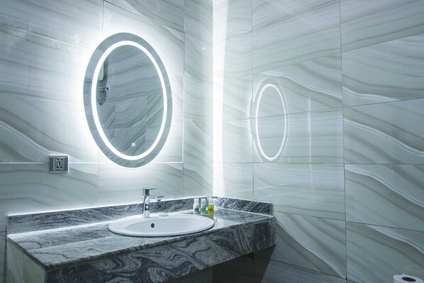 Un baño moderno con un lavamanos de mármol, un espejo grande con luz circular y una pared con diseño de mármol.