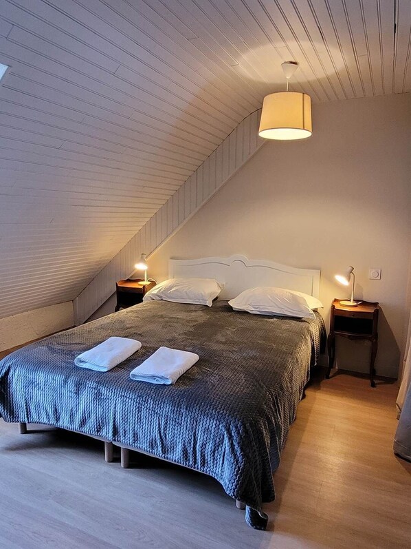 A bedroom with a sloped ceiling, a bed with a dark blanket, two bedside tables with lamps, and a hanging light fixture.