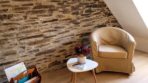 A room with a stone wall, a wooden floor, a white table with a vase of flowers, a beige armchair, and a wooden bookcase.