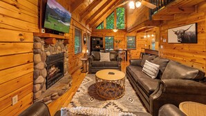 A cozy log cabin interior with a stone fireplace, leather sofas, a round coffee table, and a television mounted on the wall. - Smoky Mtn Cabin | Hot Tub | Sleep 12 | Pets | View (Gatlinburg)
