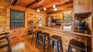 A wooden cabin interior with a kitchen, bar stools, and a large window with blinds. - Smoky Mtn Cabin | Hot Tub | Sleep 12 | Pets | View (Gatlinburg)