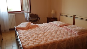 Een net opgemaakt bed met een roze sprei, een houten stoel, een klein nachtkastje met een lamp en een raam met gordijnen.