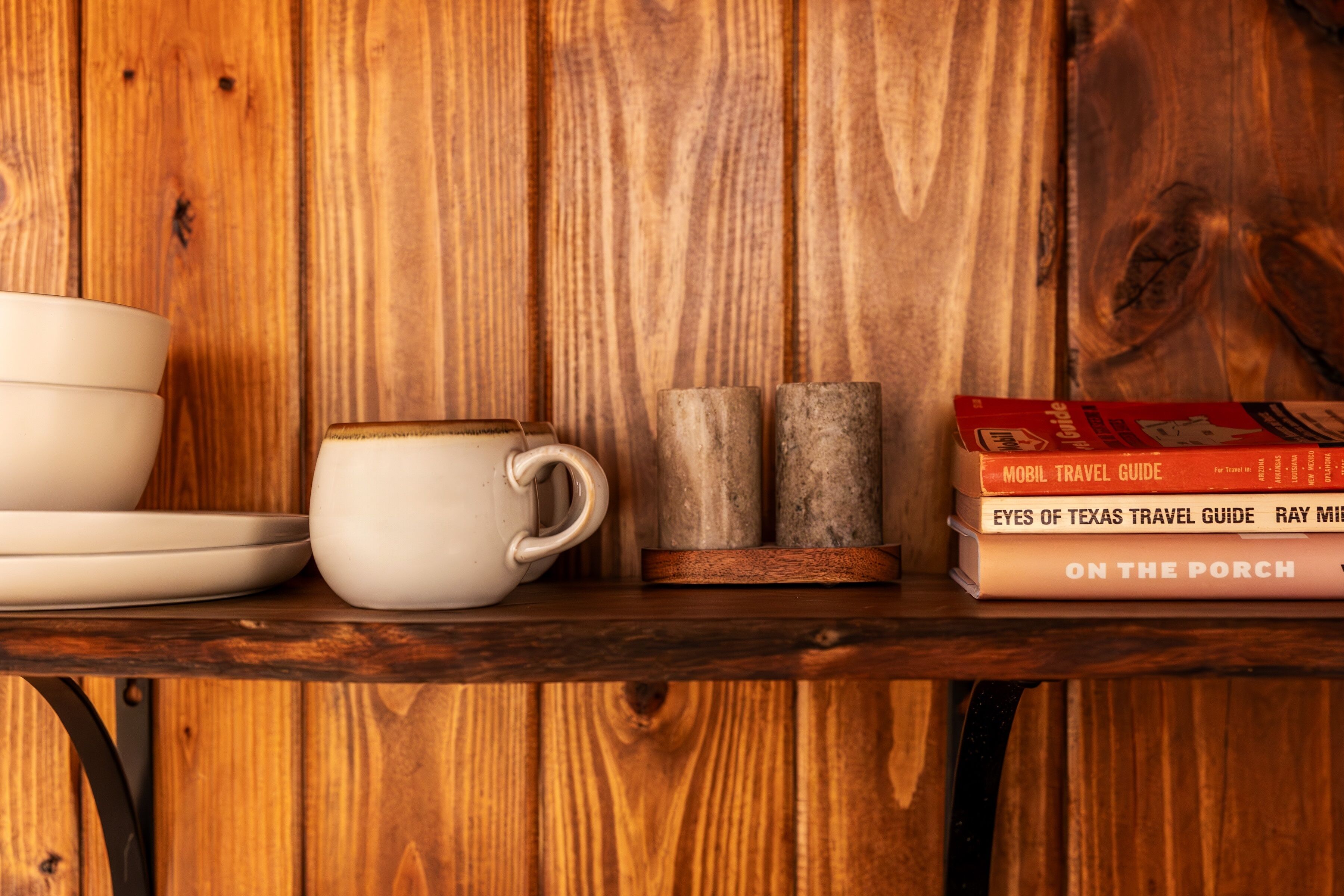 A wooden shelf with a white cup, saucer, and two rustic candle holders.