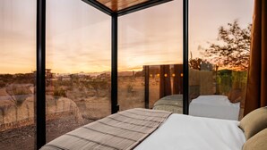 Un dormitorio con un amplio ventanal que ofrece vistas a un paisaje desértico y edificios lejanos al atardecer.