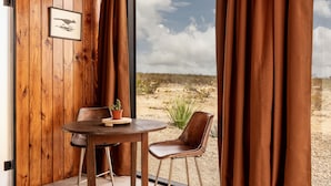 Un interior de madera con una mesa redonda y dos sillas, un cuadro enmarcado en la pared y vistas a un paisaje desértico a través de amplias ventanas.