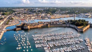 An aerial view of a harbor with numerous boats docked, a fortified structure, and a cityscape in the background.