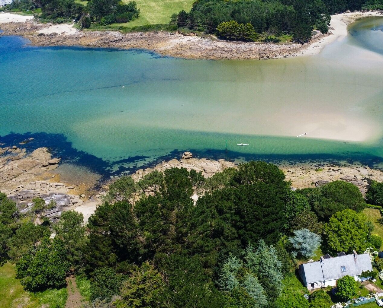 An aerial view of a coastal area with clear waters, rocky shores, dense greenery, and a small building.