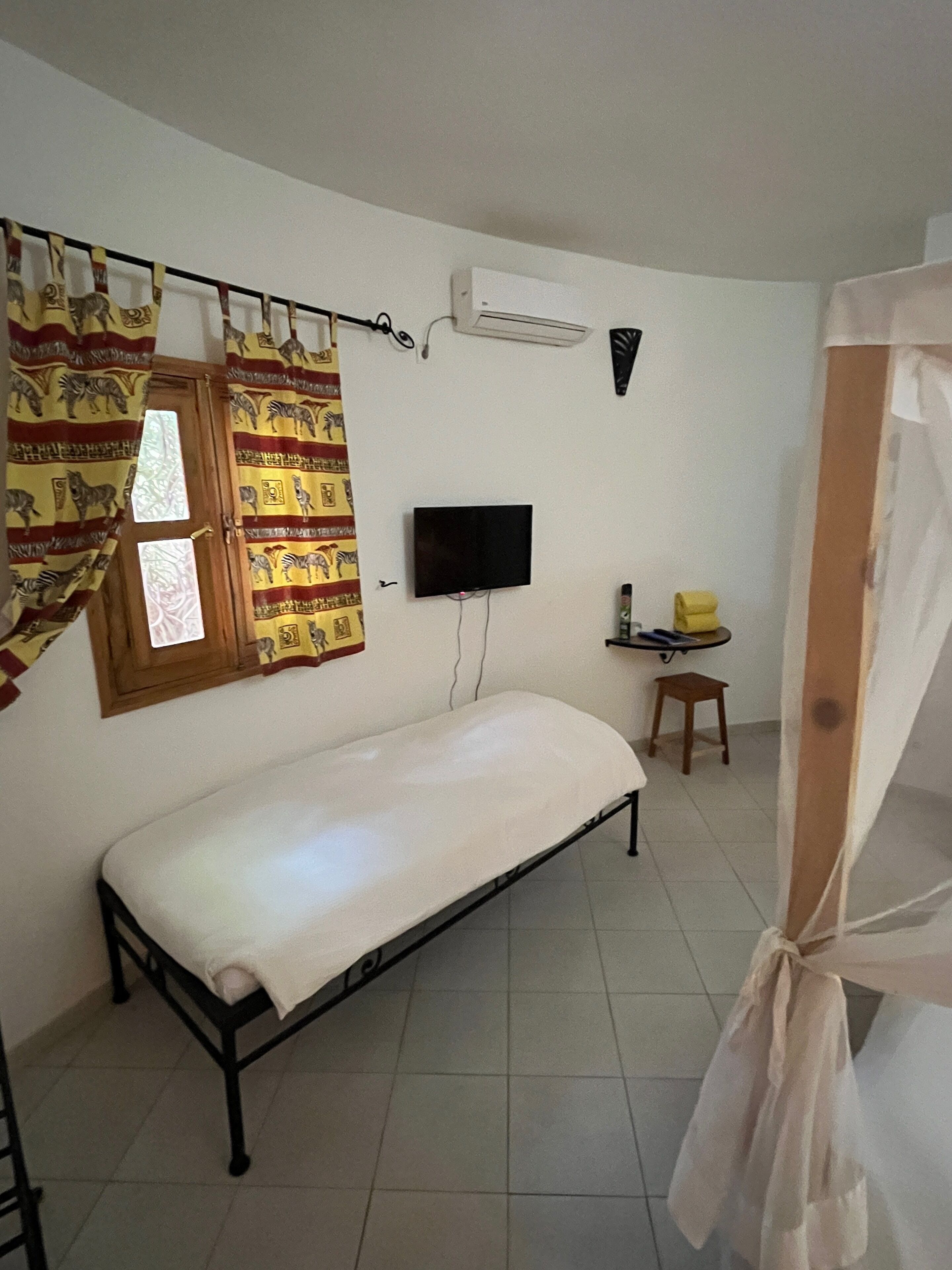 Una habitación pequeña con cama individual, televisor montado en la pared, ventana con cortinas, una mesita con una silla y un equipo de aire acondicionado.