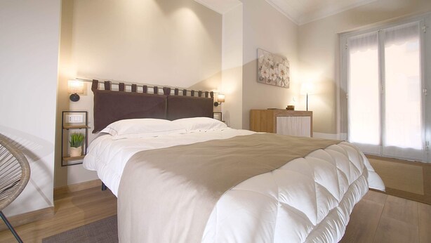 Une chambre à coucher avec un grand lit, des lampes de chevet, une petite plante et une fenêtre avec des rideaux.