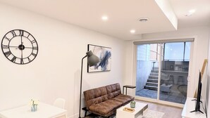 A modern living room with a white sofa, a dining table with chairs, a large clock, and a sliding glass door leading to a balcony. - Modern 2BR Suite | Private Patio | 5 Min Downtown/Zoo/Stampede | Central & Safe (Calgary)