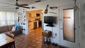 Un salon avec un canapé, une table à manger et des chaises. On aperçoit au fond une cuisine équipée d’un four à micro-ondes et de placards. Un ventilateur de plafond est visible, et un téléviseur est fixé au mur.