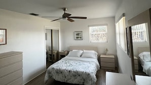 Une chambre à coucher comprenant un lit, une commode, une table de chevet, un ventilateur de plafond et une fenêtre donnant sur un paysage agréable.