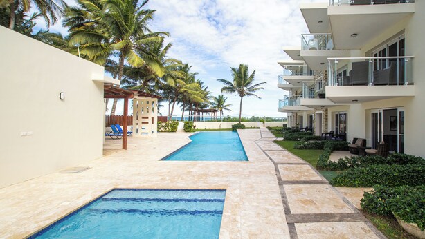 A swimming pool area with a clear view of the pool, palm trees, and a building with balconies. - 0047Cozy 1 bedroom Ocean View  Cabarete (Cabarete)