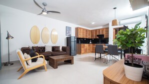 A modern living room with a brown sofa, wooden coffee table, and a dining area with a wooden table and chairs. A ceiling fan and a potted plant are visible. - 0047Cozy 1 bedroom Ocean View  Cabarete (Cabarete)