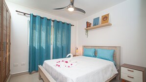 A bedroom with a bed, a nightstand, a ceiling fan, a window with curtains, and a shelf with pictures. - 0047Cozy 1 bedroom Ocean View  Cabarete (Cabarete)