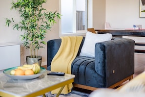 Un salon moderne avec un canapé foncé, un plaid jaune, une plante en pot et un panier de fruits posé sur une table.