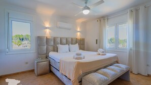Un dormitori amb llit, taules de nit, una banca, un ventilador de sostre i finestres amb cortines.