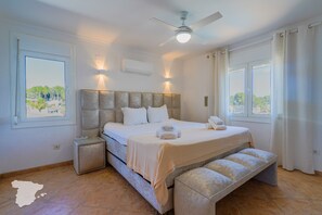A bedroom with a bed, bedside tables, a bench, a ceiling fan, and windows with curtains.
