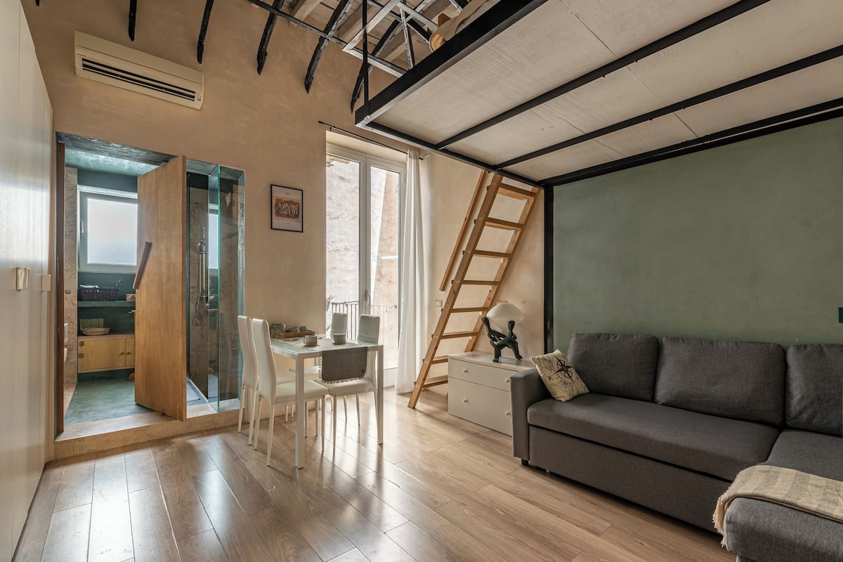 A modern living room with a sofa, a dining table with chairs, a ladder, and a view of the outside.