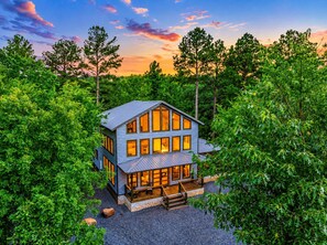 A two-story house surrounded by dense greenery, with a visible deck and large windows. - Playset & Pool Table | Hot Tub | Sleeps 14 (Broken Bow)