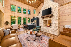 A spacious living area with a stone fireplace, wooden walls, and a large window overlooking greenery. - Playset & Pool Table | Hot Tub | Sleeps 14 (Broken Bow)