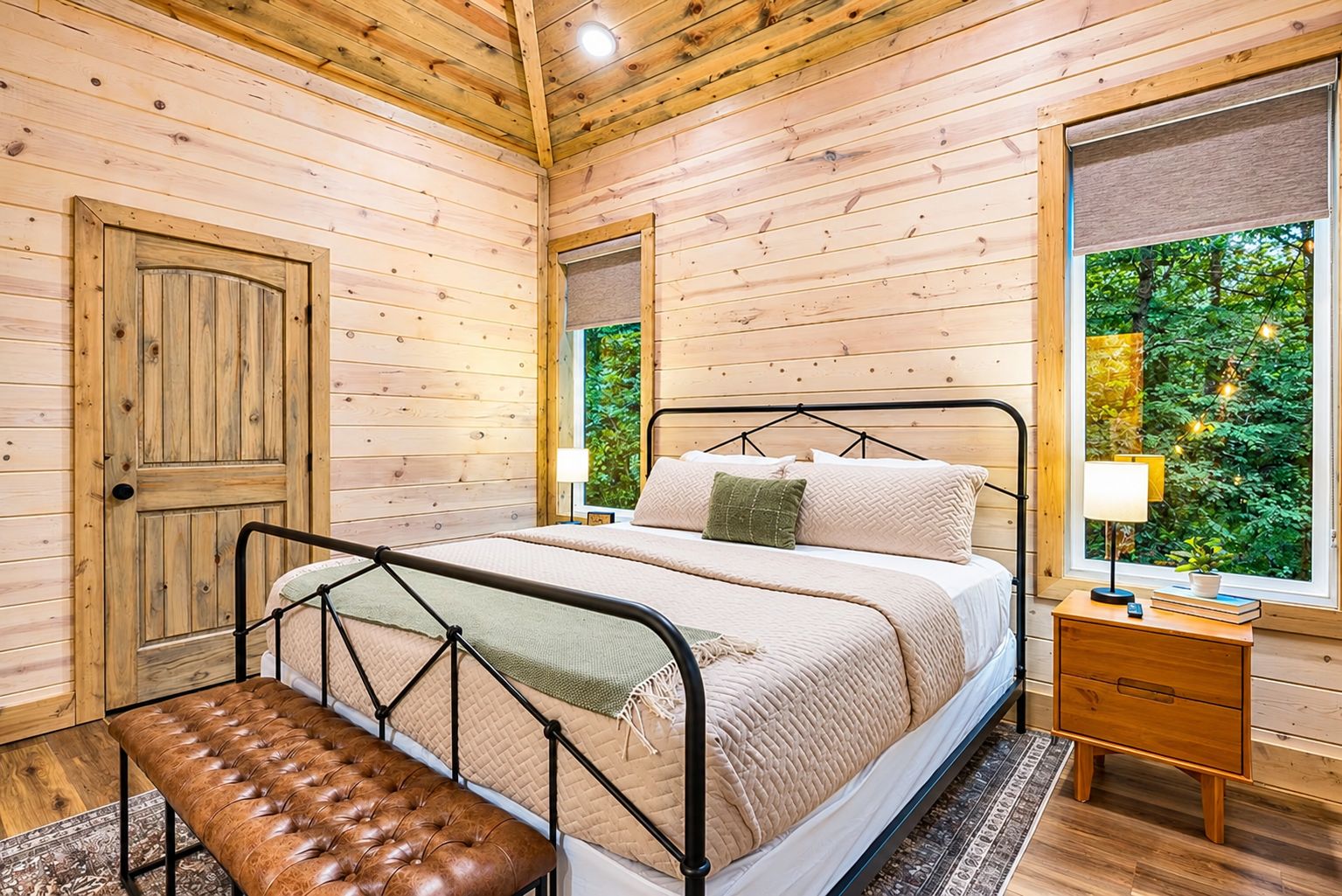 Un dormitorio de cabaña de madera con cama, banco, mesita de noche y ventana con vistas a la vegetación.