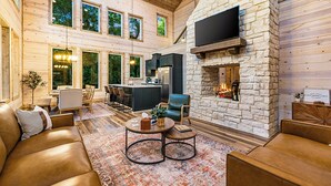 A spacious living area with a stone fireplace, wooden walls, and a large TV mounted on the wall. - 3BR Forest Hideaway! Playset, Pool Table, Foosball (Broken Bow)