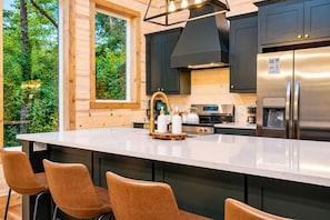 A modern kitchen with a large island, dark cabinetry, stainless steel appliances, and a view of greenery outside. - Playset & Pool Table | Hot Tub | Sleeps 14 (Broken Bow)