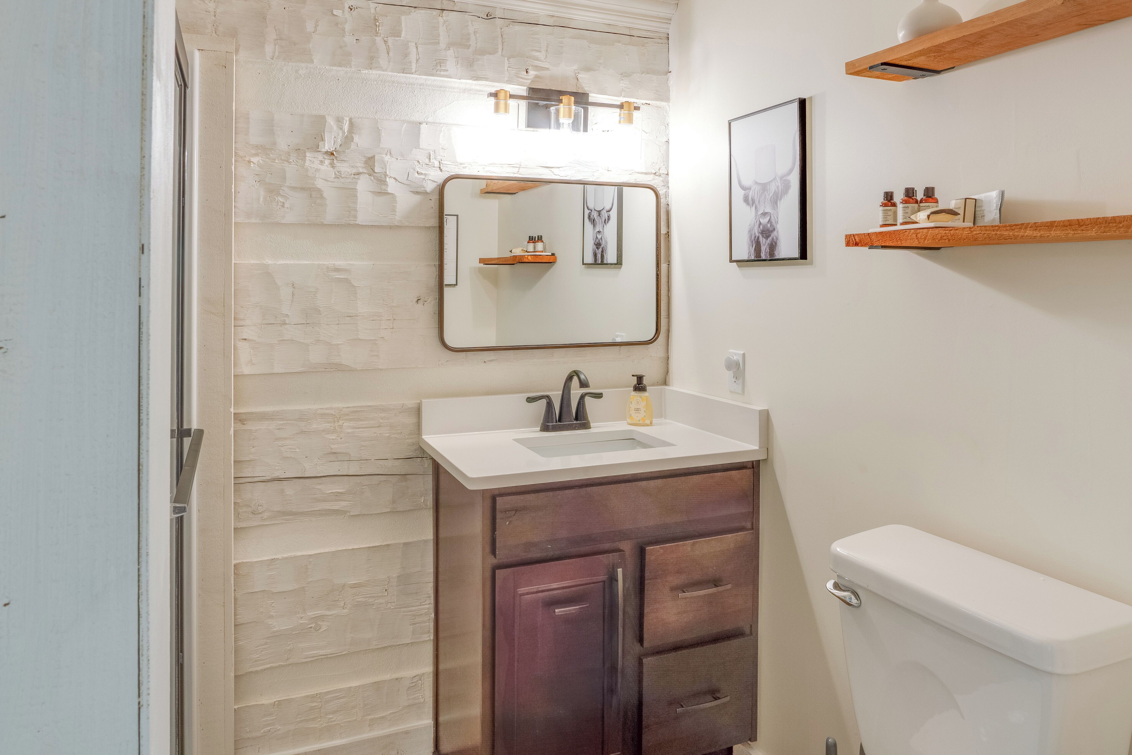 Une salle de bain avec un lavabo blanc, des meubles en bois foncé, des toilettes blanches et un miroir surmonté d’une étagère en bois.