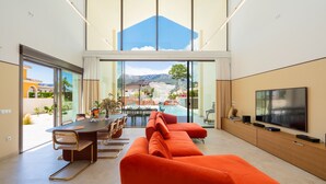 Uma sala de estar moderna com uma televisão grande, um sofá, um espaço de refeições com cadeiras e vista para uma paisagem montanhosa através de janelas amplas.