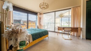 Quarto com cama, escrivaninha, cadeira, janela ampla e vista para edifícios.