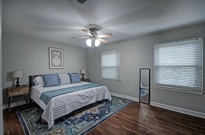 Cozy retreat with a large bed, nightstand, and mirror, set against a backdrop of a ceiling fan and window with blinds.