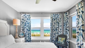 A bedroom with a large bed, a nightstand with a lamp, a chair with a cushion, and a view of the beach through the window. - Emerald Dunes 103 (Destin)