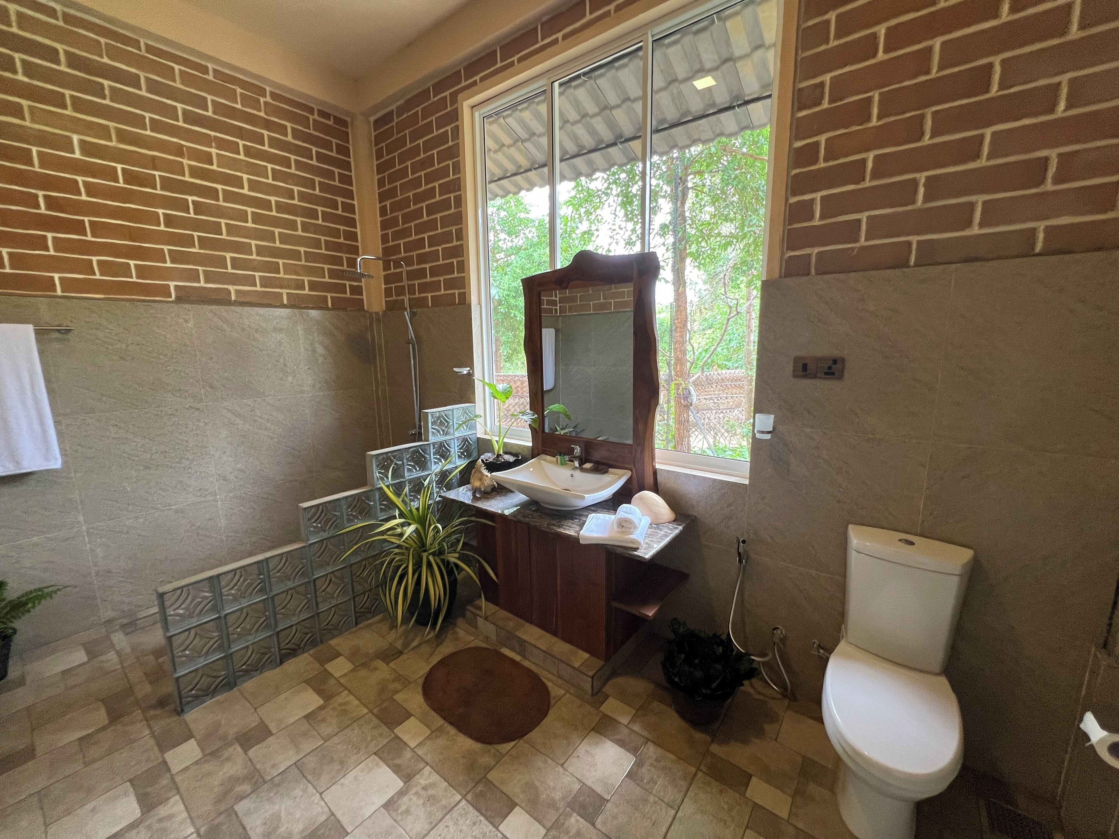 Un baño con piso de baldosas, un lavamanos con espejo, un inodoro y una ventana con vistas a follaje.