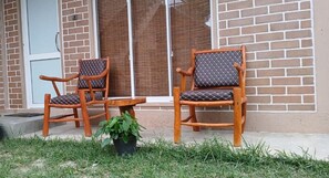 Two wooden chairs with cushioned seats and backs, a small table, and a potted plant on a concrete patio in front of a brick building with a sliding glass door.