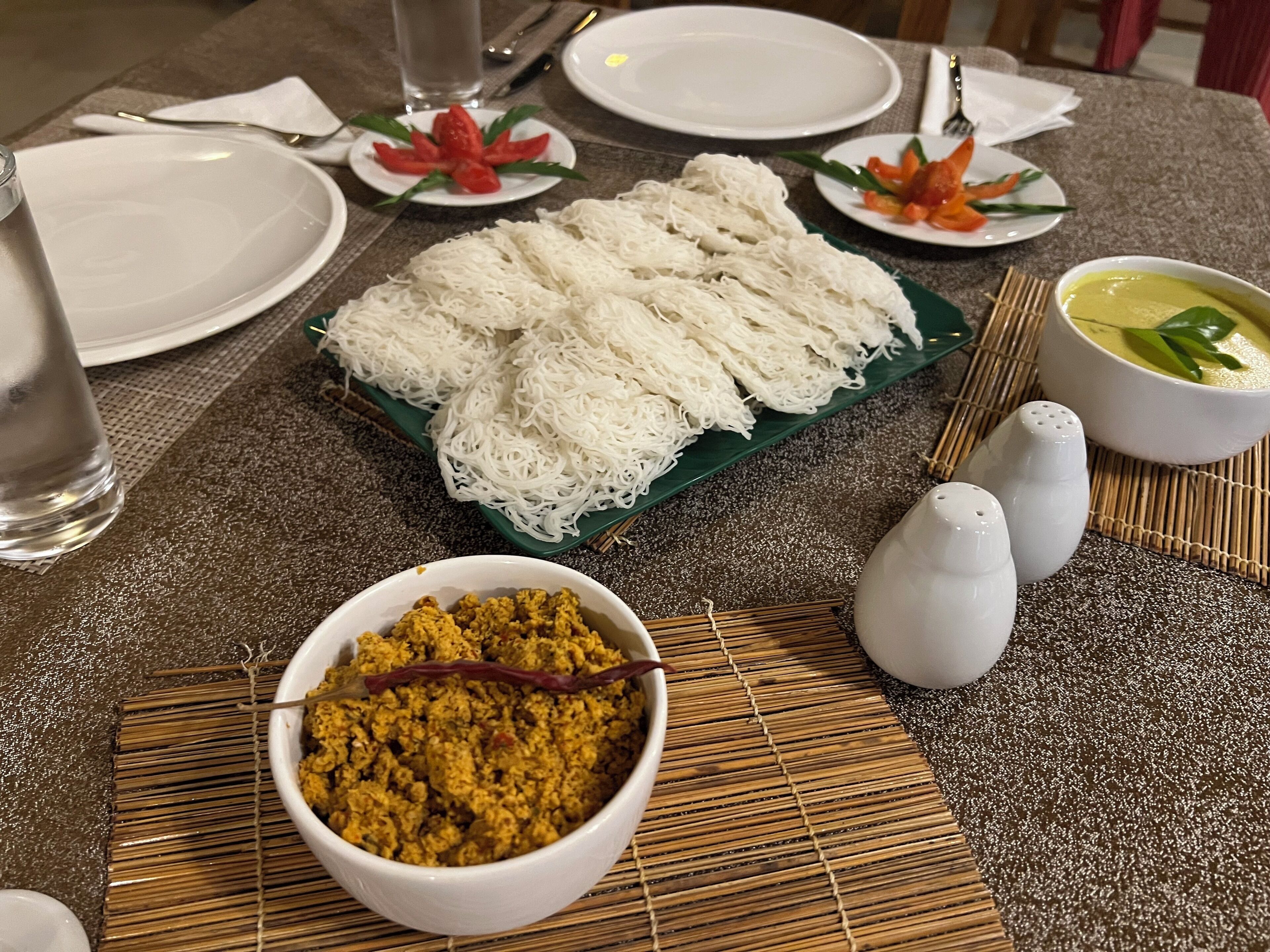 A table set with white plates, a bowl of yellow curry, a bowl of white noodles, and a bowl of green curry.