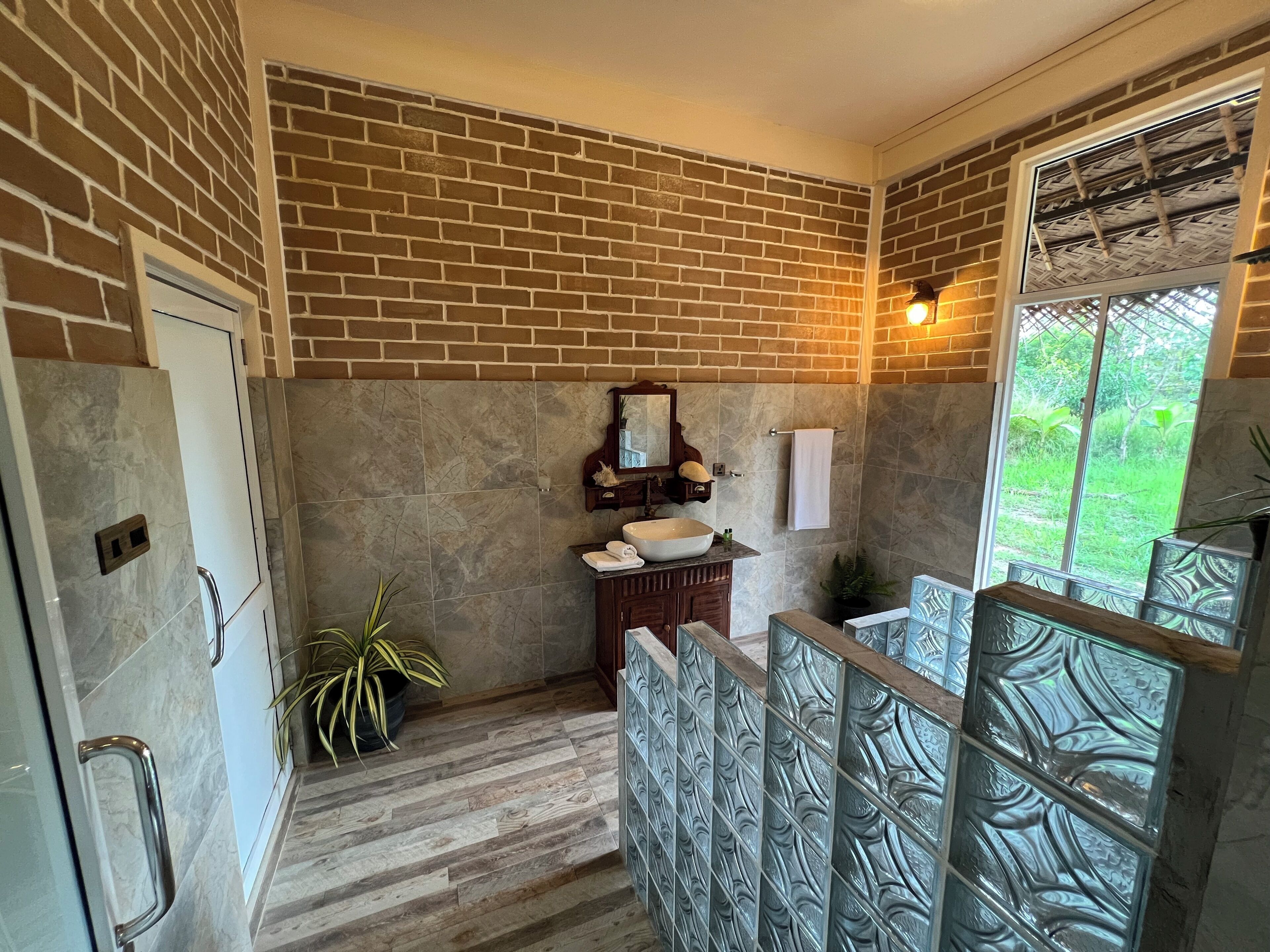 Un baño con pared de ladrillo, tocador de madera, un espejo grande y una ventana con vistas a la vegetación.